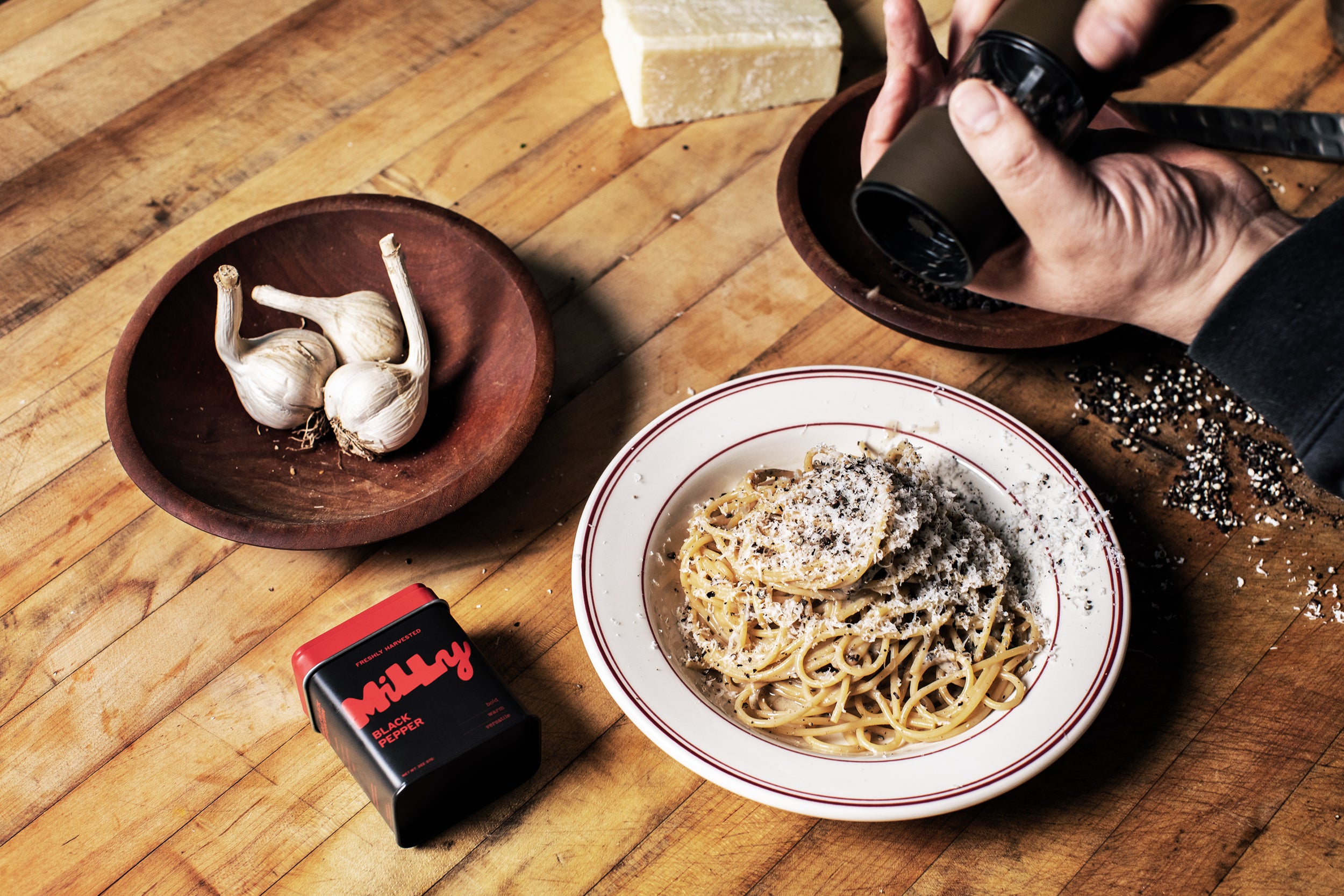 Person seasoning spaghetti with a black container on a wooden table with garlic and cheese.