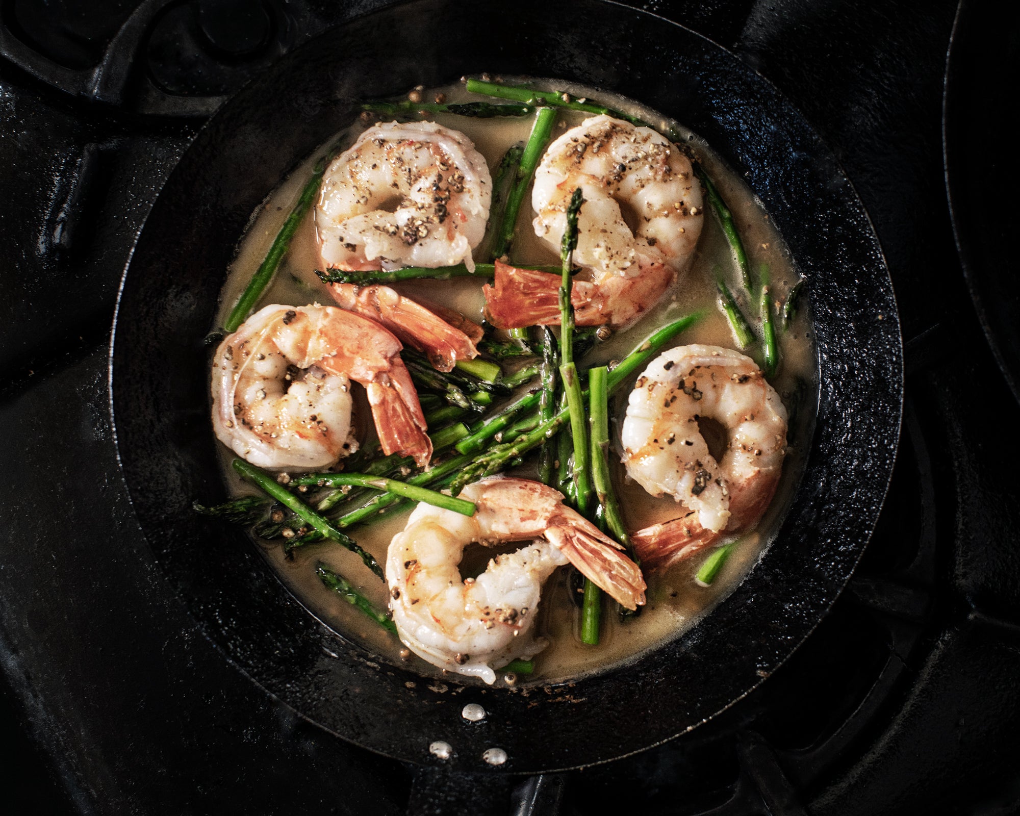 A top-down view of shrimp and asparagus cooking in a black cast iron skillet on a dark stovetop.
