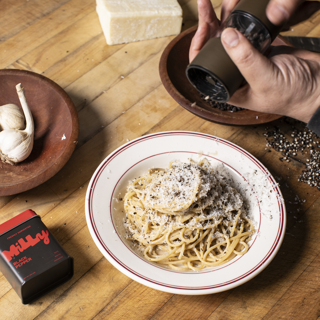 A hand using a matte black Milly pepper grinder to finish freshly ground black pepper over a bowl of cacio e pepe pasta on a wooden table, with a Milly Black Pepper tin, garlic, and Parmesan nearby.