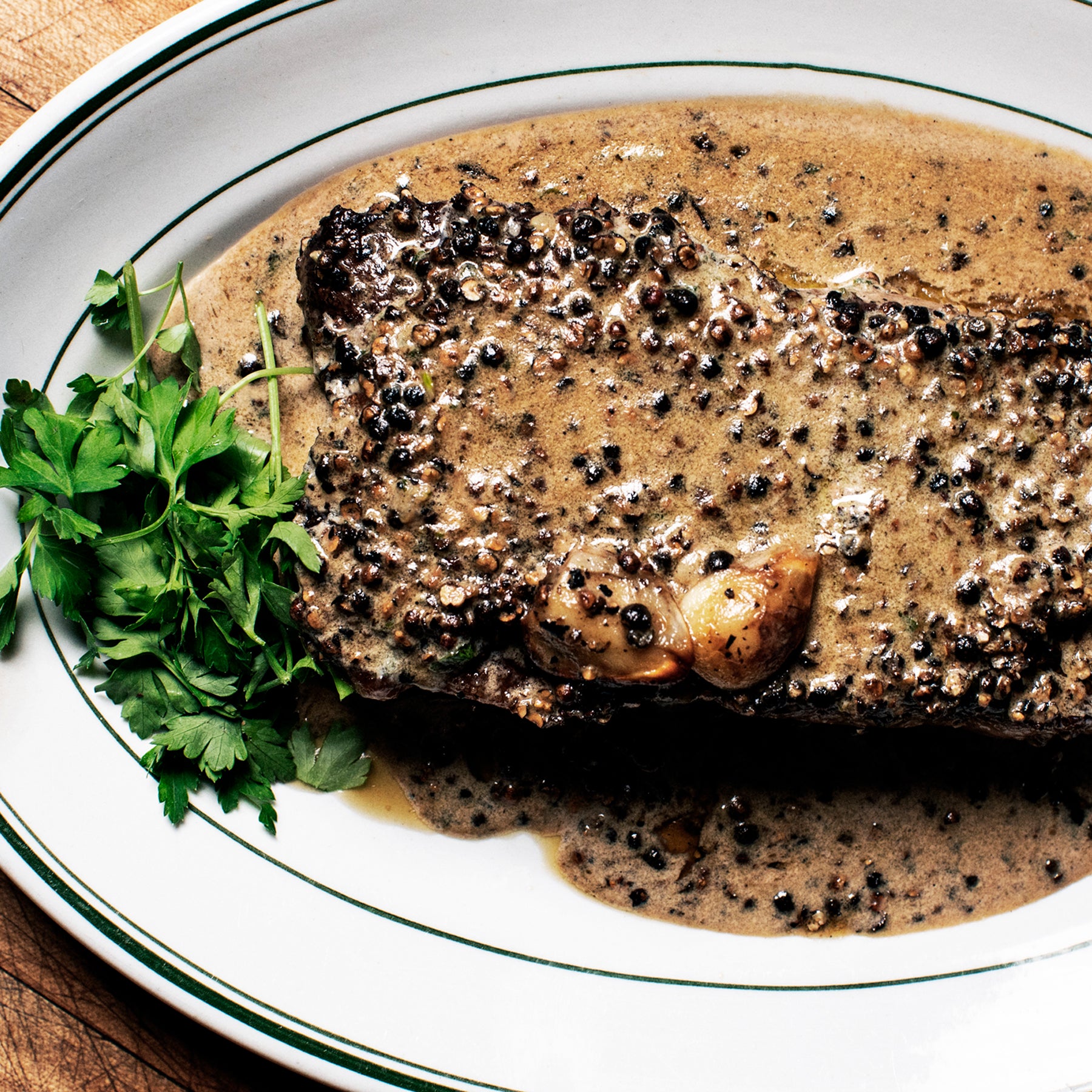 A plated steak au poivre on a white plate with green-striped rim, coated in creamy peppercorn sauce and garnished with fresh parsley leaves.