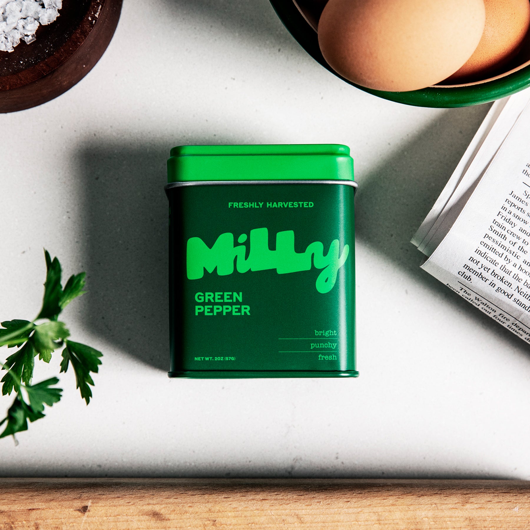 A dark green Milly Green Pepper tin on a gray marble surface surrounded by fresh parsley, brown eggs in a bowl, and an open book, evoking a cozy kitchen setting.