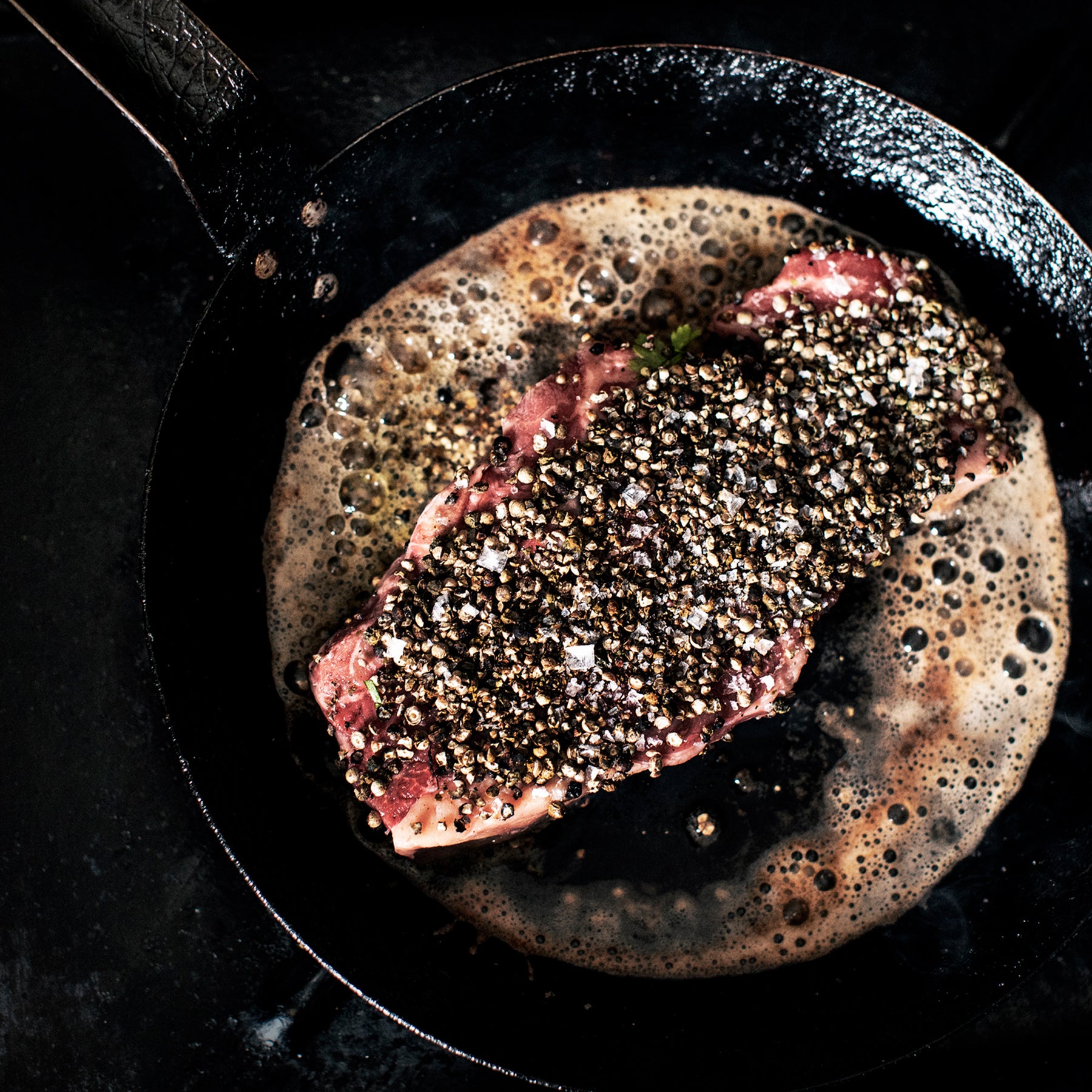 A pepper-crusted raw steak sizzling in a cast iron skillet with foamy butter, shot from directly above on a dark background.